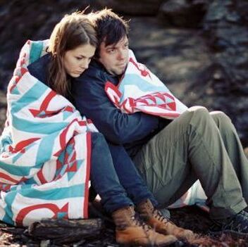 Man and Woman sit huddled under a quilt